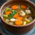 Crock pot chicken broth - the image is a close-up of a bowl of soup. the bowl is made of ceramic and has a dark brown color with a speckled pattern. the soup appears to be a broth with chunks of chicken, carrots, celery, and other vegetables mixed in. there is a sprig of parsley on top of the soup, adding a pop of green color to the dish. the background is a dark wooden table.