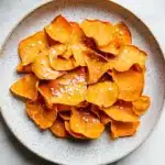 Dehydrated sweet potato chips air fryer - the image shows a plate of orange-colored potato chips on a white marble countertop. the chips are arranged in a circular pattern on the plate, with some overlapping each other. they are sprinkled with sesame seeds, giving them a crunchy texture. in the top right corner of the image, there is an air fryer, suggesting that the chips are being prepared for cooking.