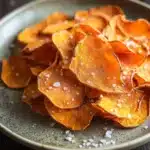 Dehydrated sweet potato chips air fryer - the image shows a plate of sweet potato chips on a dark wooden table. the chips are arranged in a circular pattern on the plate, with some overlapping each other. they are golden brown in color and have a crispy texture. there are small white salt sprinkled on top of the chips, adding a pop of color to the dish. in the background, there are more chips scattered around the plate. the overall mood of the image is rustic and appetizing.