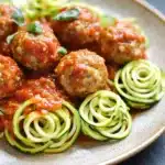 Dinner turkey meatballs with zucchini noodles - the image shows a plate of spaghetti and meatballs. the plate is made of light-colored ceramic and has a beige rim. the meatballs are covered in a red tomato sauce and are arranged in a circular pattern on top of the spaghetti noodles. there are also some fresh basil leaves scattered around the plate. the spaghetti noodles are thin and spiral-like, and they are arranged around the meatballs in a way that they are overlapping each other. the dish appears to be freshly cooked and ready to eat.