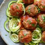 Dinner turkey meatballs with zucchini noodles - the image shows a plate of zucchini noodles with meatballs. the noodles are cooked al dente and are arranged in a circular pattern on the plate. the meatballs are covered in a red tomato sauce and garnished with chopped parsley. the plate is white with a speckled pattern and is sitting on a dark grey countertop.