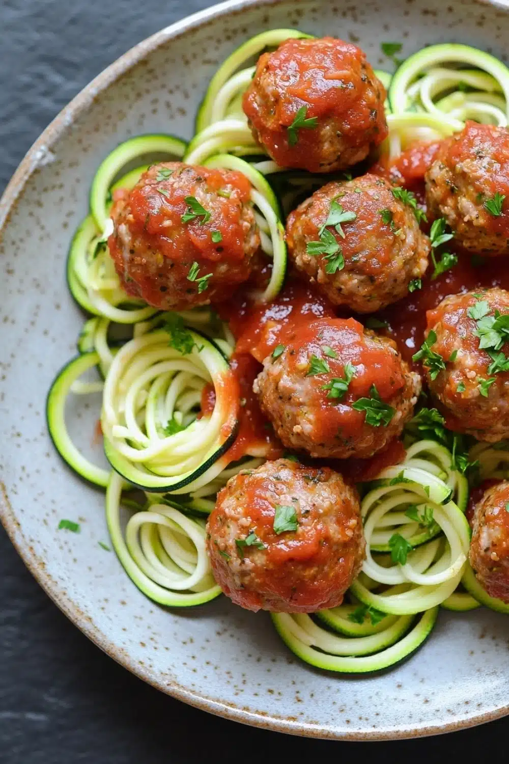 Dinner turkey meatballs with zucchini noodles - the image shows a plate of zucchini noodles with meatballs. the noodles are cooked al dente and are arranged in a circular pattern on the plate. the meatballs are covered in a red tomato sauce and garnished with chopped parsley. the plate is white with a speckled pattern and is sitting on a dark grey countertop.