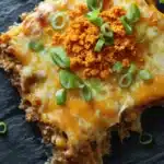 Dorito casserole freezer meal - the image shows a slice of lasagna on a black slate plate. the lasagna has a golden brown crust on top and is topped with melted cheese and chopped green onions. the cheese is melted and bubbly, and there is a layer of ground beef on top. the dish appears to be freshly baked and ready to eat. the plate is garnished with fresh green onions, adding a pop of color to the dish.
