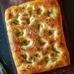 Focaccia bread - the image shows a rectangular piece of bread on a wooden cutting board. the bread appears to be freshly baked and has a golden brown crust. it is topped with a sprinkle of white sesame seeds and fresh rosemary sprigs. the cutting board is placed on a dark grey countertop with a green cloth napkin on the side. the overall aesthetic of the image is rustic and natural.