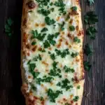 Garlic bread with pizza dough - the image shows a rectangular piece of bread with a golden brown crust on a dark wooden surface. the bread appears to be freshly baked and has a layer of melted cheese on top. the cheese is white and bubbly, and there are small chunks of meat scattered throughout the cheese. on top of the cheese, there is a sprig of fresh parsley, adding a pop of green color to the dish. the parsley is scattered around the bread, creating a contrast with the dark wood. the overall aesthetic of the image is rustic and appetizing.