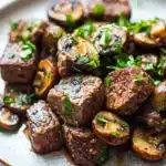 Garlic butter steak bites with mushrooms crockpot - the image is a close-up of a plate of food. the plate is white with a speckled pattern and is placed on a wooden table. on the plate, there are several pieces of cooked meat, which appear to be beef tenderloin, arranged in a circular pattern. the meat is dark brown in color and has a marbled texture. there are also small pieces of sliced mushrooms scattered around the plate. the mushrooms are also garnished with chopped parsley, adding a pop of green color to the dish. the dish looks appetizing and ready to eat.