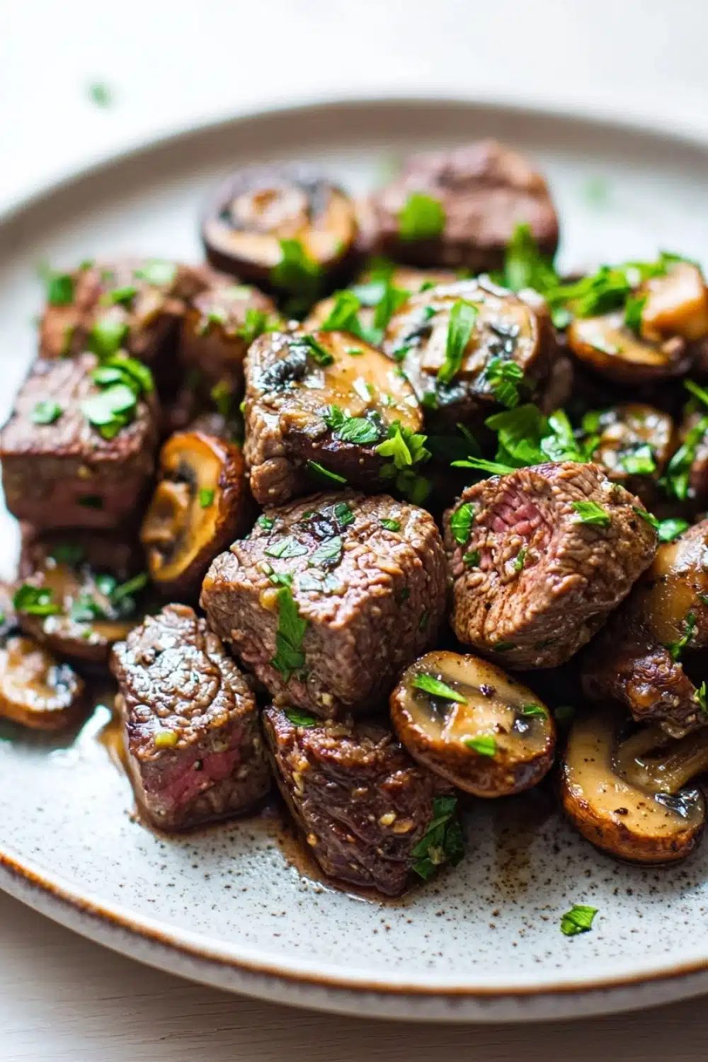 Garlic butter steak bites with mushrooms crockpot - the image is a close-up of a plate of food. the plate is white with a speckled pattern and is placed on a wooden table. on the plate, there are several pieces of cooked meat, which appear to be beef tenderloin, arranged in a circular pattern. the meat is dark brown in color and has a marbled texture. there are also small pieces of sliced mushrooms scattered around the plate. the mushrooms are also garnished with chopped parsley, adding a pop of green color to the dish. the dish looks appetizing and ready to eat.