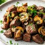 Garlic butter steak bites with mushrooms crockpot - the image is a close-up of a plate of food. the plate is white with a speckled pattern and is placed on a white marble countertop. on top of the plate, there are several pieces of cooked beef, which appear to be medium-rare and have a reddish-brown color. the beef is garnished with sliced mushrooms and chopped parsley. the mushrooms are arranged in a pile and are scattered around the plate. the dish looks appetizing and ready to eat.