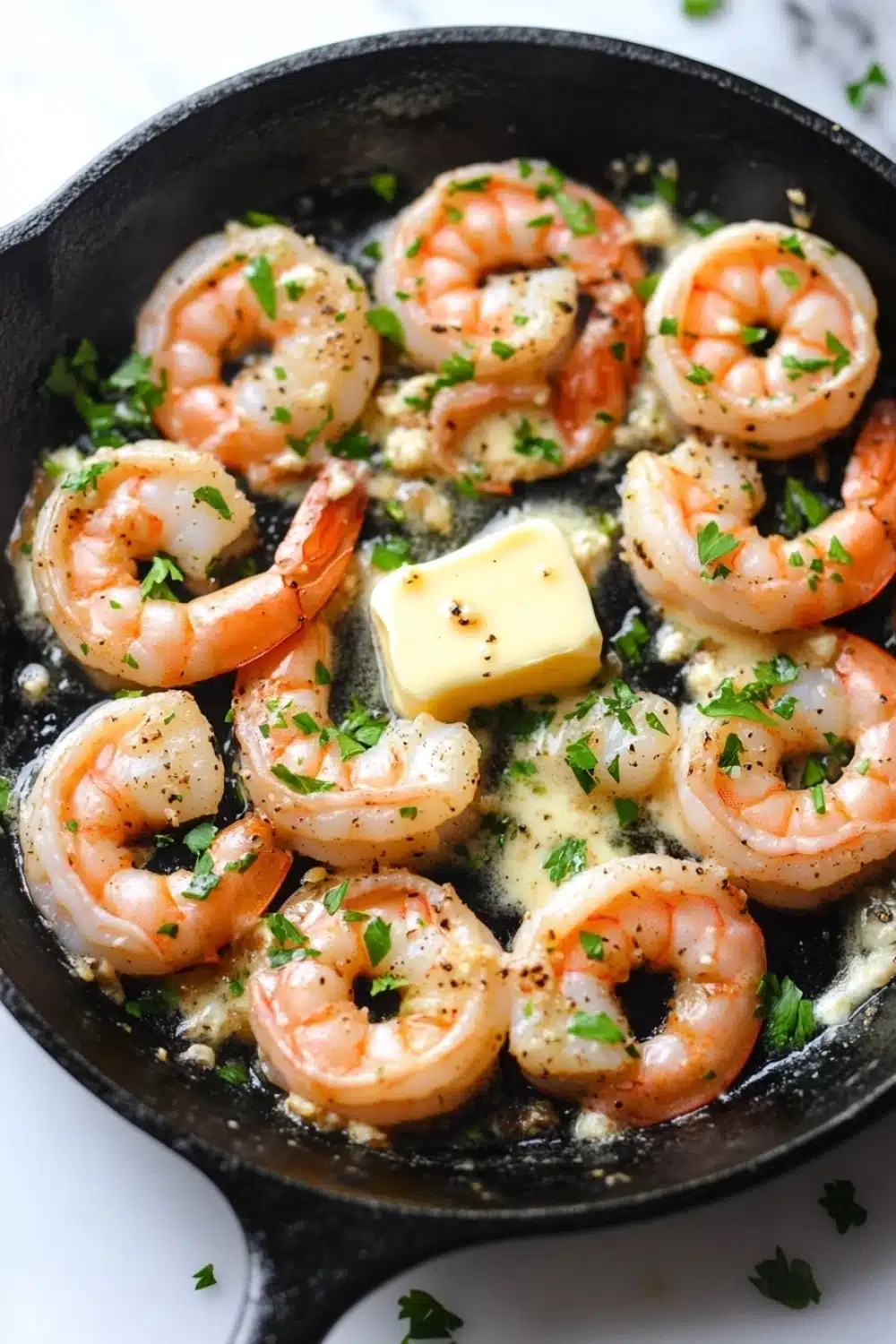 Garlic shrimp in cast iron skillet - the image shows a black cast iron skillet with cooked shrimp in it. the shrimp are pink and appear to be seasoned with herbs and spices. they are arranged in a single layer in the skillet, with some of them overlapping each other. there is a small block of butter on top of the shrimp, which is yellow and has flecks of herbs scattered throughout. the skillet is sitting on a white marble countertop.