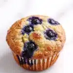 Gluten free lemon blueberry muffin - the image is of a freshly baked blueberry muffin on a white marble surface. the muffin has a golden brown crust and is filled with fresh blueberries. the blueberries are arranged in a circular pattern on top of the muffin, with some of them overlapping each other. the edges of the blueberries have a crumbly texture, indicating that they have been freshly baked. the background is blurred, making the muffins the focal point of the image.