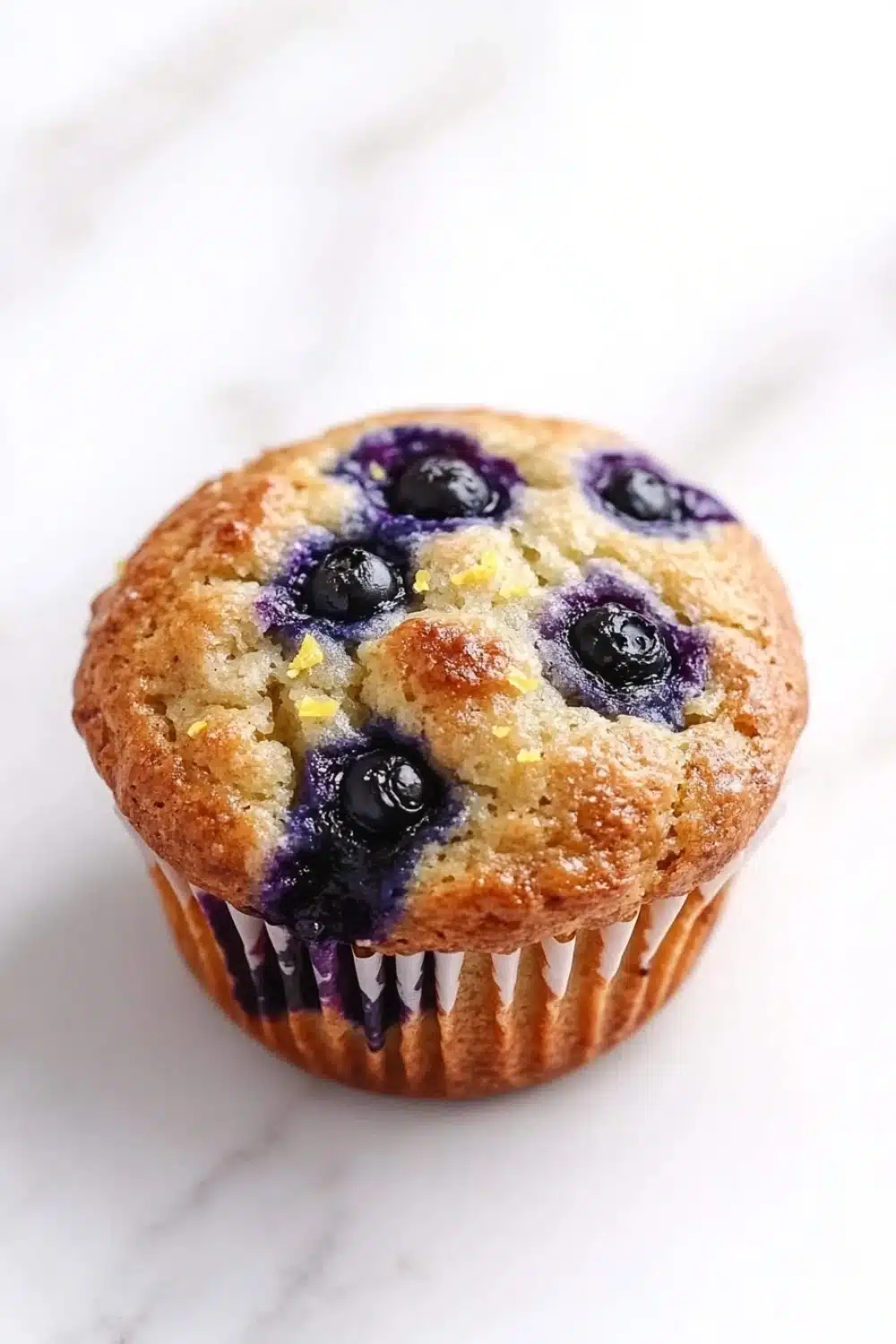Gluten free lemon blueberry muffin - the image is of a freshly baked blueberry muffin on a white marble surface. the muffin has a golden brown crust and is filled with fresh blueberries. the blueberries are arranged in a circular pattern on top of the muffin, with some of them overlapping each other. the edges of the blueberries have a crumbly texture, indicating that they have been freshly baked. the background is blurred, making the muffins the focal point of the image.