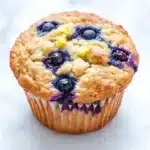 Gluten free lemon blueberry muffin - the image is of a freshly baked blueberry muffin on a white marble surface. the muffin has a golden brown crust on top and is filled with fresh blueberries. the blueberries are arranged in a circular pattern around the edges of the muffin, with some of them overlapping each other. there is a crumbly topping on top, which appears to be a mixture of blueberries and yellow flowers. the background is blurred, making the blueberries the focal point of the image.