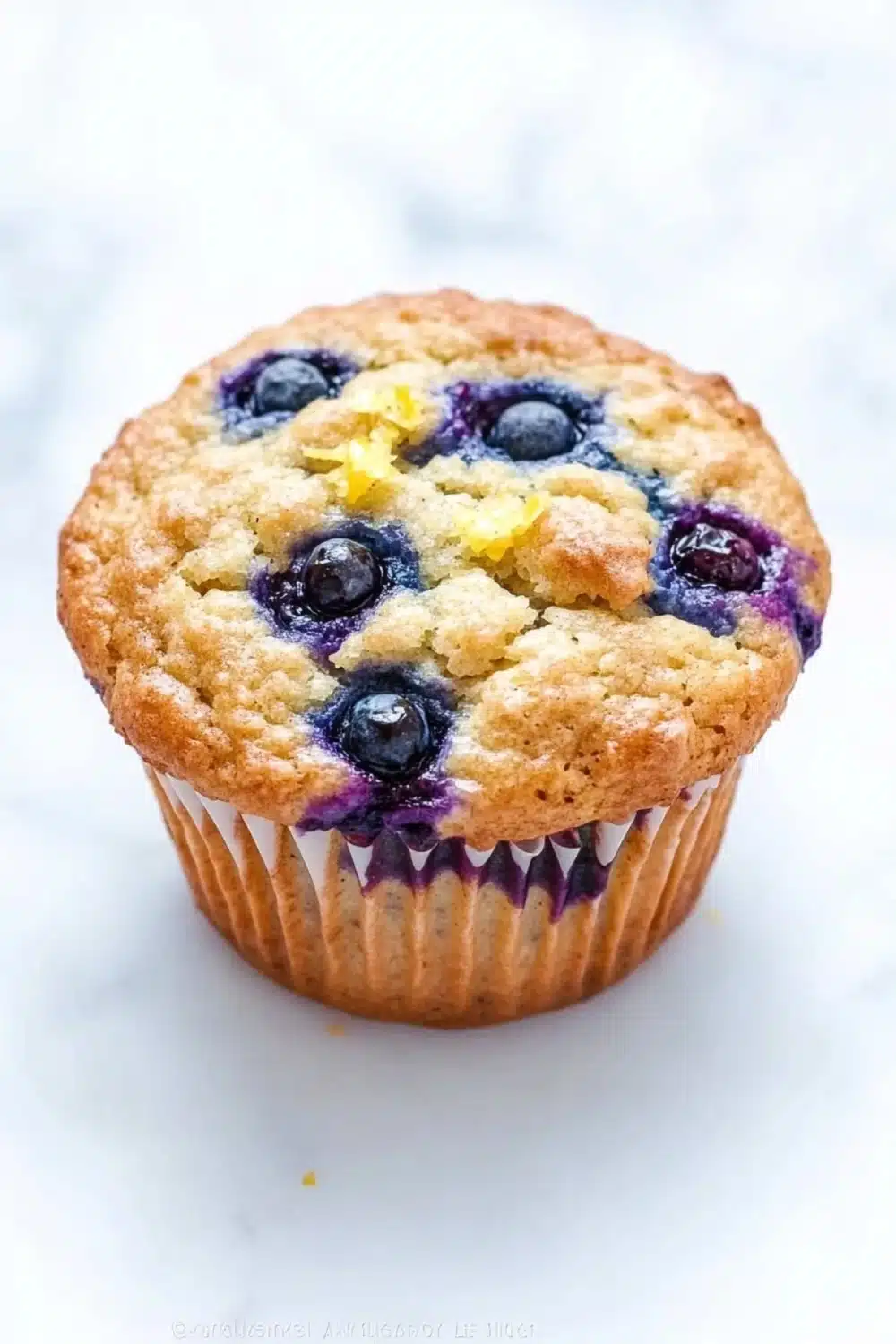 Gluten free lemon blueberry muffin - the image is of a freshly baked blueberry muffin on a white marble surface. the muffin has a golden brown crust on top and is filled with fresh blueberries. the blueberries are arranged in a circular pattern around the edges of the muffin, with some of them overlapping each other. there is a crumbly topping on top, which appears to be a mixture of blueberries and yellow flowers. the background is blurred, making the blueberries the focal point of the image.