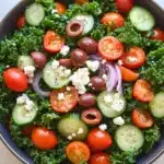 Greek salad kale - the image is a close-up of a salad in a blue bowl. the salad is made up of kale leaves, sliced cucumbers, cherry tomatoes, olives, and feta cheese. the tomatoes are bright red and appear to be ripe and juicy. the cucumbers are cut into thin slices and are arranged in a circular pattern. the olives are scattered throughout the salad, adding a pop of color and texture. the feta is crumbled and adds a creamy texture to the dish. the bowl is sitting on a light blue countertop.
