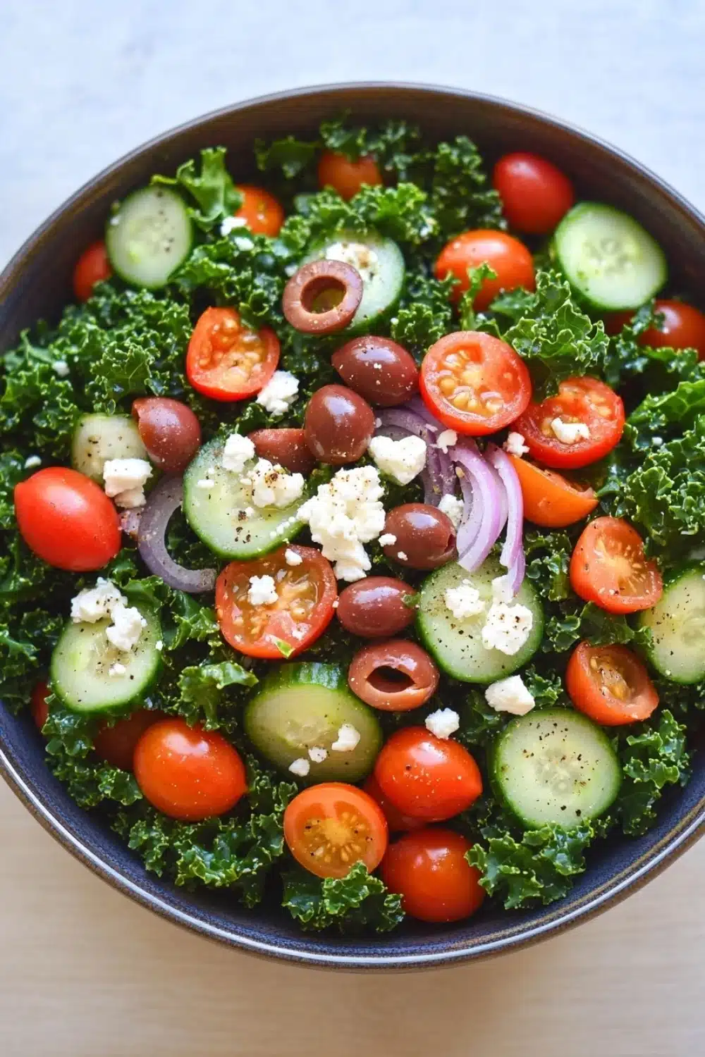 Greek salad kale - the image is a close-up of a salad in a blue bowl. the salad is made up of kale leaves, sliced cucumbers, cherry tomatoes, olives, and feta cheese. the tomatoes are bright red and appear to be ripe and juicy. the cucumbers are cut into thin slices and are arranged in a circular pattern. the olives are scattered throughout the salad, adding a pop of color and texture. the feta is crumbled and adds a creamy texture to the dish. the bowl is sitting on a light blue countertop.