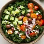 Greek salad kale - the image is of a large wooden bowl filled with a colorful salad. the salad is made up of kale leaves, sliced cucumbers, cherry tomatoes, red onions, and feta cheese. the tomatoes are bright red and appear to be ripe and juicy. the cucumbers are cut into small cubes and are scattered throughout the salad. there are also some olives and red onions scattered throughout. the bowl is sitting on a white wooden table with a sprig of parsley on the side. the overall color scheme of the salad is vibrant and appetizing.