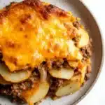 Ground beef and sliced potato casserole - the image is a close-up of a plate of food. the plate is round and has a light brown color with a speckled pattern. on top of the plate, there is a layer of melted cheese that is golden brown and bubbly. the cheese appears to be melted and oozing out of the sides. below the cheese, there are chunks of ground beef, diced potatoes, and onions. the dish looks freshly baked and ready to eat.
