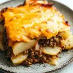 Ground beef and sliced potato casserole - the image shows a slice of lasagna on a gray plate. the lasagna has a golden brown crust on top and is covered in melted cheese. the cheese is melted and bubbly, and it appears to be freshly baked. on top of the lasagna, there is a layer of ground beef and diced potatoes. the plate is sitting on a white marble countertop.