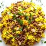 Ground beef and yellow rice for dinner - the image shows a close-up of a dish made with yellow rice and ground beef. the rice appears to be cooked and has a golden-brown color. the ground beef is scattered throughout the dish, with some pieces of diced onions and chopped parsley scattered around it. the dish is served on a white parchment paper. the background is blurred, making the dish the focal point of the image.