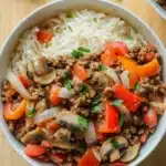 Ground beef dinner make ahead - the image shows a bowl of food on a wooden table. the bowl is white and has a serving of white rice on top. the food appears to be a dish made with ground beef, sliced mushrooms, red bell peppers, and onions. there are also some fresh basil leaves scattered around the bowl. in the background, there is another bowl of the same dish. the overall color scheme of the image is warm and inviting.