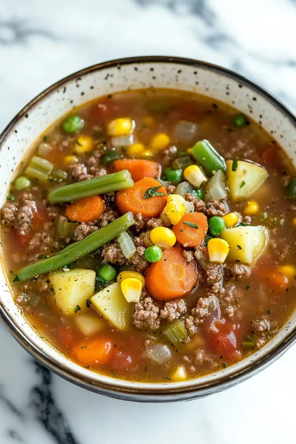 Ground beef for dinner soup - the image is a close-up of a bowl of soup. the soup appears to be a vegetable soup with chunks of ground beef, carrots, corn, and green onions mixed in. the bowl is made of ceramic and has a speckled pattern on the rim. the background is a white marble countertop.