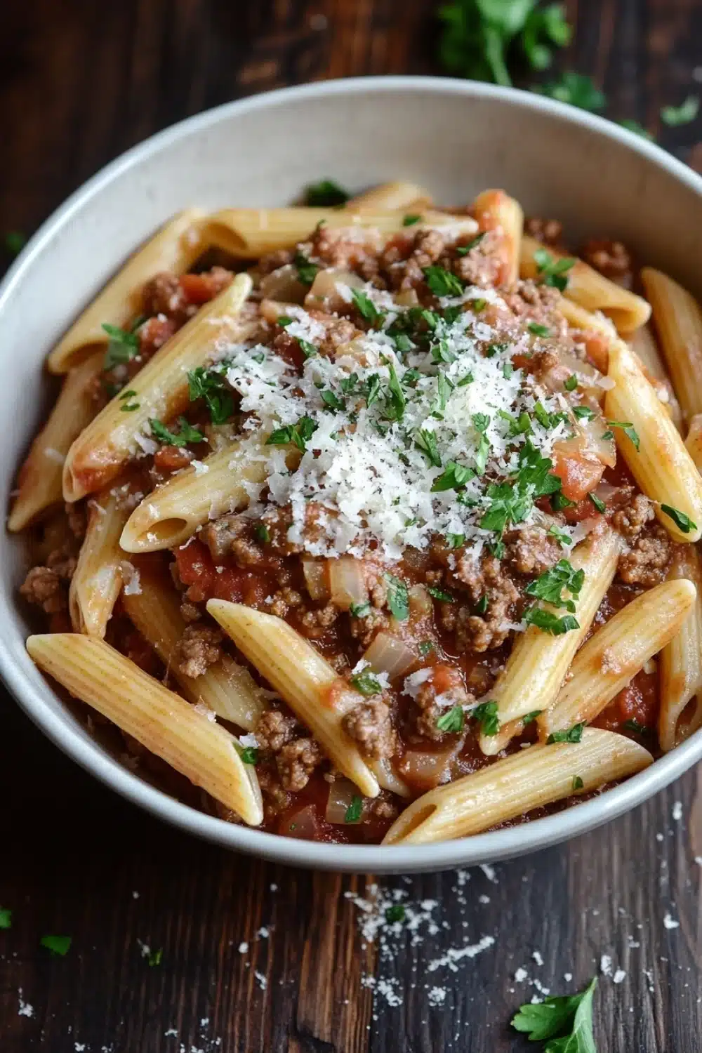 Ground beef pasta dish - the image is a close-up of a bowl of penne pasta dish. the pasta is cooked al dente and is topped with a rich, red sauce and chunks of ground meat. the sauce appears to be tomato-based and is drizzled over the top. the dish is garnished with chopped parsley and grated parmesan cheese. the bowl is sitting on a wooden table with a few sprigs of parsley scattered around it. the overall color palette of the dish is warm and inviting.