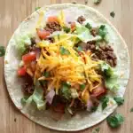 Ground beef skillet taco - the image is of a taco on a wooden table. the taco is made with a tortilla shell and is filled with ground beef, diced tomatoes, onions, and cilantro. on top of the beef, there is a generous amount of shredded cheese. the cheese is yellow and appears to be melted and bubbly. there are also a few sprigs of cilantro scattered around the tortilla, adding a pop of green color to the dish.