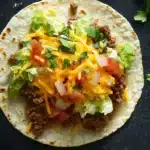 Ground beef skillet taco - the image is a close-up of a taco on a black surface. the taco is made with a tortilla shell and is filled with ground beef, diced tomatoes, onions, and cilantro. the cheese is melted and bubbly on top, and there are two lime wedges on either side of the tortilla. there are also a few sprigs of cilantro scattered around the dish. the background is dark, making the taco stand out.