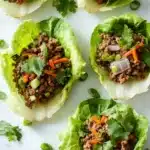 High protein beef lettuce wraps - the image shows four lettuce wraps filled with ground beef and vegetables. the lettuce wraps are arranged on a white marble surface and are garnished with fresh cilantro. the ground beef appears to be cooked and is mixed with diced carrots, onions, and green onions. the vegetables are bright orange and appear to be seasoned with herbs and spices. there are also a few sprigs of parsley scattered around the lettuce wraps.