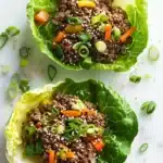 High protein beef lettuce wraps - the image shows two lettuce wraps filled with a mixture of ground beef and vegetables. the lettuce wraps are placed on a white marble countertop with scattered green onions and sesame seeds around them. the ground beef appears to be cooked and has a brown color, while the vegetables are a mix of orange, yellow, and green colors. the dish looks appetizing and ready to eat.
