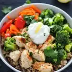 High protein chicken and rice bowl - the image is a close-up of a bowl of food. the bowl is white and is placed on a black countertop. the food in the bowl appears to be a stir-fry dish with rice, chicken, broccoli, carrots, and bell peppers. there is a dollop of sour cream on top of the rice. a lemon wedge is placed next to the bowl, and there are a few sprigs of cilantro scattered around the bowl. the overall color scheme of the dish is warm and inviting.