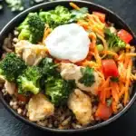 High protein chicken and rice bowl - the image is a close-up of a bowl of food. the bowl is black and is placed on a black slate surface. the food appears to be a stir-fry dish with rice, broccoli, carrots, and chicken. there is a dollop of sour cream on top of the rice. the broccoli is bright green and looks fresh, while the carrots are bright orange. the chicken is cut into small pieces and is scattered throughout the dish. there are also some cilantro leaves scattered around the bowl, adding a pop of green color.