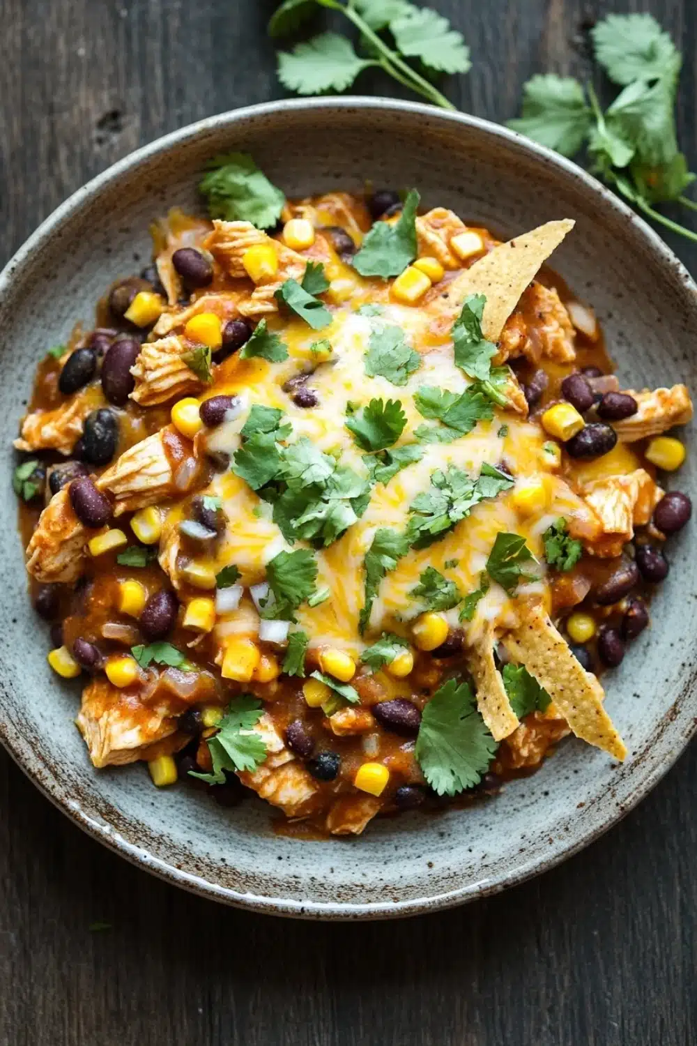 High protein chicken enchilada skillet - the image is a close-up of a bowl of chili. the chili is made with black beans, corn kernels, and shredded chicken. the chicken is cooked to a golden brown color and appears to be seasoned with herbs and spices. there are also a few tortilla chips scattered on top of the chili. a sprig of cilantro is also visible in the background. the bowl is sitting on a wooden table.