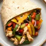 High protein chicken fajita burritos - the image shows a tortilla wrap on a blue plate. the wrap is filled with shredded chicken, diced bell peppers, black beans, and red onions. the chicken is cooked to a golden brown color and appears to be seasoned with herbs and spices. the bell peppers are bright red and yellow, while the black beans are black. the cheese is melted and bubbly on top of the wrap. the plate is sitting on a white marble countertop.