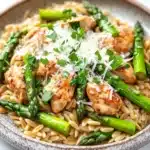 High protein chicken orzo with asparagus - the image is a close-up of a plate of pasta dish. the pasta is fusilli, which is a type of spiral-shaped pasta, and is topped with grilled chicken and asparagus. the chicken is cooked to a golden brown color and appears to be seasoned with herbs and spices. the dish is garnished with a sprinkle of parmesan cheese. the plate is sitting on a white marble countertop.