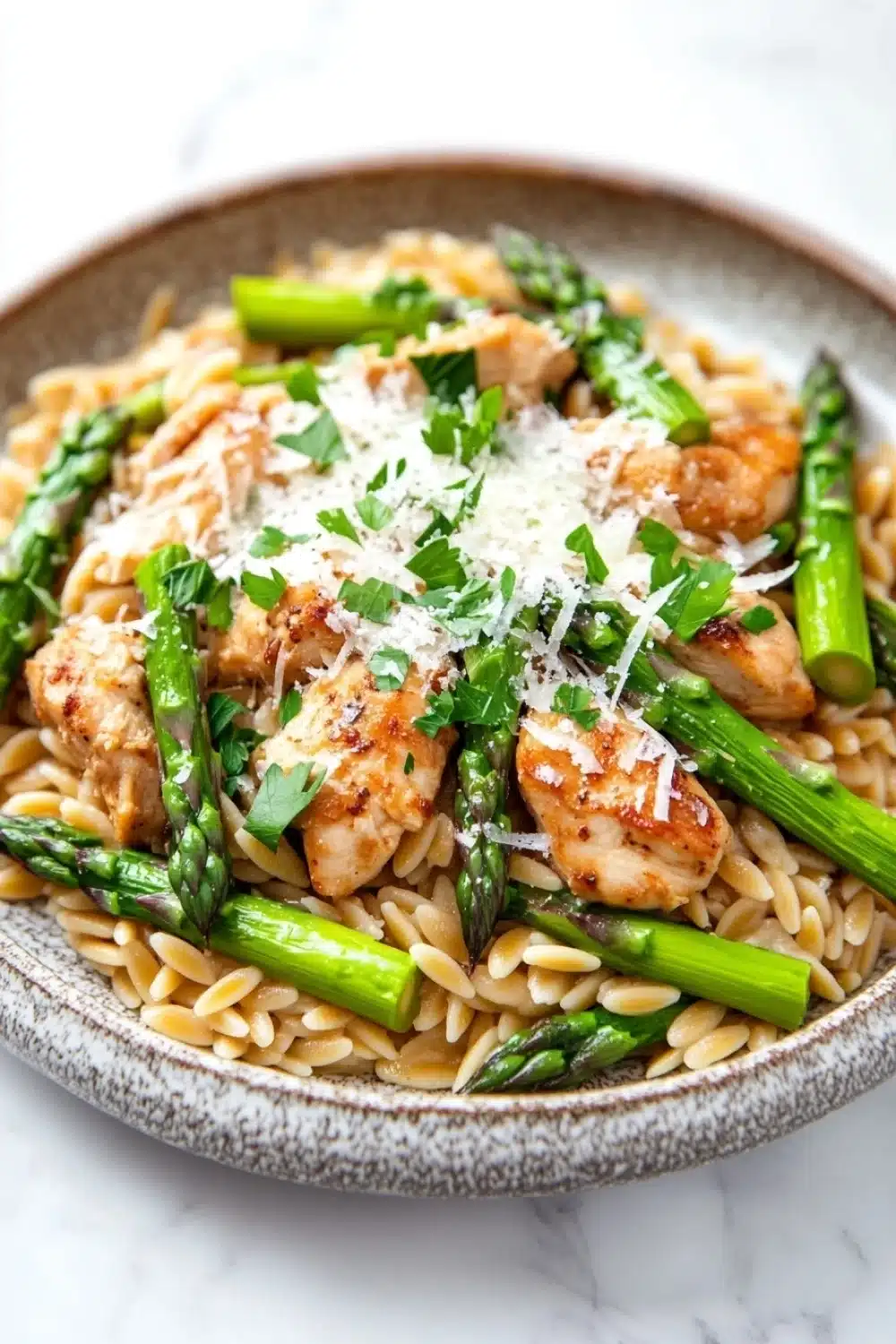 High protein chicken orzo with asparagus - the image is a close-up of a plate of pasta dish. the pasta is fusilli, which is a type of spiral-shaped pasta, and is topped with grilled chicken and asparagus. the chicken is cooked to a golden brown color and appears to be seasoned with herbs and spices. the dish is garnished with a sprinkle of parmesan cheese. the plate is sitting on a white marble countertop.