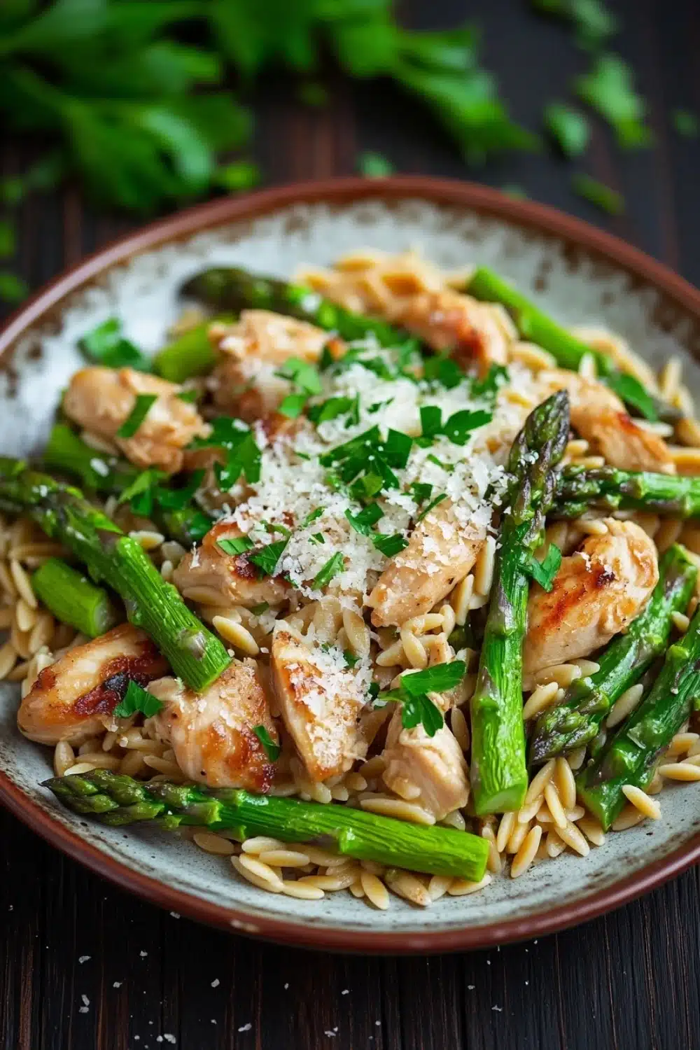 High protein chicken orzo with asparagus - the image is a close-up of a plate of pasta dish. the pasta appears to be fusilli, which is a type of spiral pasta, and is topped with grilled chicken and asparagus. the chicken is cooked to a golden brown color and is garnished with chopped parmesan cheese and fresh herbs. the dish is served on a dark wooden table with a few sprigs of parsley scattered around. the overall color palette of the dish is warm and inviting.