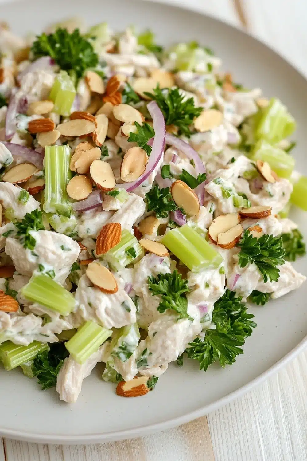 High protein chicken salad with greek yogurt - the image is a close-up of a plate of salad. the salad appears to be made with chicken, celery, red onion, and almonds. the chicken is white and fluffy, and the celery is cut into small pieces. the almonds are scattered throughout the salad, adding a crunchy texture and nutty flavor. the celery and onions are bright green and look fresh and vibrant. the plate is white, and it is sitting on a white wooden table.