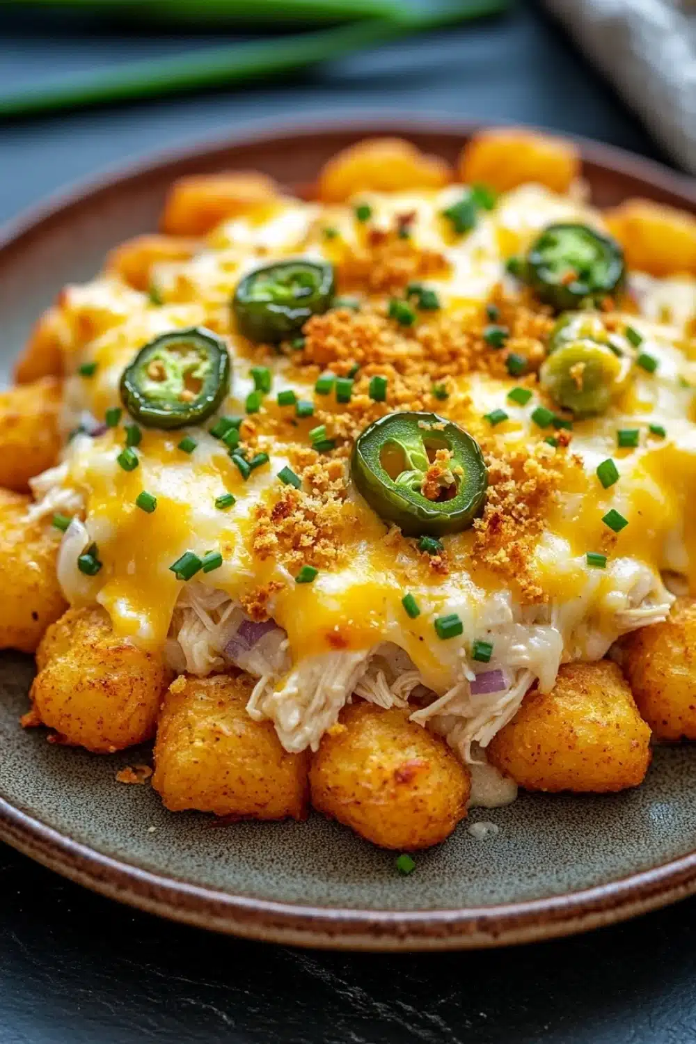 Jalapeno popper tater tot chicken casserole - the image shows a plate of food, specifically a casserole dish. the dish appears to be made with chicken, cheese, and jalapeno peppers. the chicken is cooked to a golden brown color and is covered in melted cheese. the cheese is melted and bubbly, and there are small pieces of green onions scattered throughout the dish. on top of the chicken, there is a sprinkle of breadcrumbs. the plate is brown and has a dark brown rim. the background is blurred, but it seems to be a kitchen countertop with a few green onions and a white napkin.