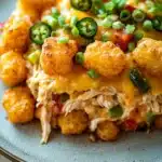 Jalapeno popper tater tot chicken casserole - the image is a close-up of a plate of food. the plate is blue and has a speckled pattern. on top of the plate, there is a serving of shredded chicken with a layer of melted cheese on top. the chicken is golden brown and appears to be seasoned with herbs and spices. there are also small pieces of green jalapeno peppers scattered throughout the dish. the dish looks appetizing and ready to eat.