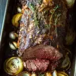 Keto slow cooker roast beef - the image shows a large piece of cooked beef tenderloin in a rectangular baking dish. the beef is cooked medium-rare and has a dark brown crust on top. it is garnished with a sprig of fresh thyme. the dish is served on a bed of sliced onions, which are arranged in a circular pattern around the roast. the onions are caramelized and have a golden-brown color, indicating that they have been cooked to perfection. the roast is drizzled with a dark sauce, which is visible on the sides of the dish. there are also a few small pieces of garlic scattered around the dish, adding a pop of green color to the image. the baking dish is sitting on a wooden table.