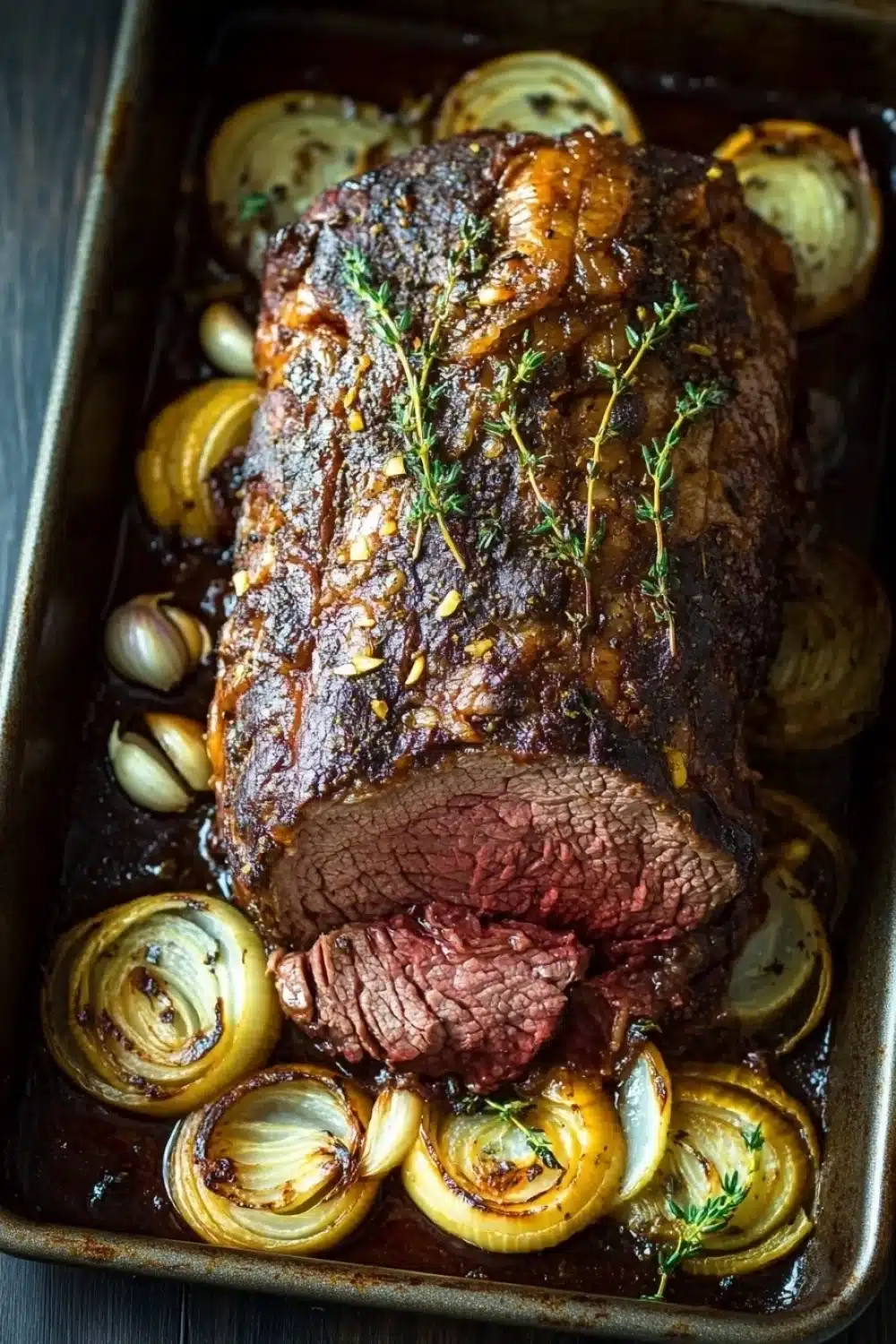 Keto slow cooker roast beef - the image shows a large piece of cooked beef tenderloin in a rectangular baking dish. the beef is cooked medium-rare and has a dark brown crust on top. it is garnished with a sprig of fresh thyme. the dish is served on a bed of sliced onions, which are arranged in a circular pattern around the roast. the onions are caramelized and have a golden-brown color, indicating that they have been cooked to perfection. the roast is drizzled with a dark sauce, which is visible on the sides of the dish. there are also a few small pieces of garlic scattered around the dish, adding a pop of green color to the image. the baking dish is sitting on a wooden table.