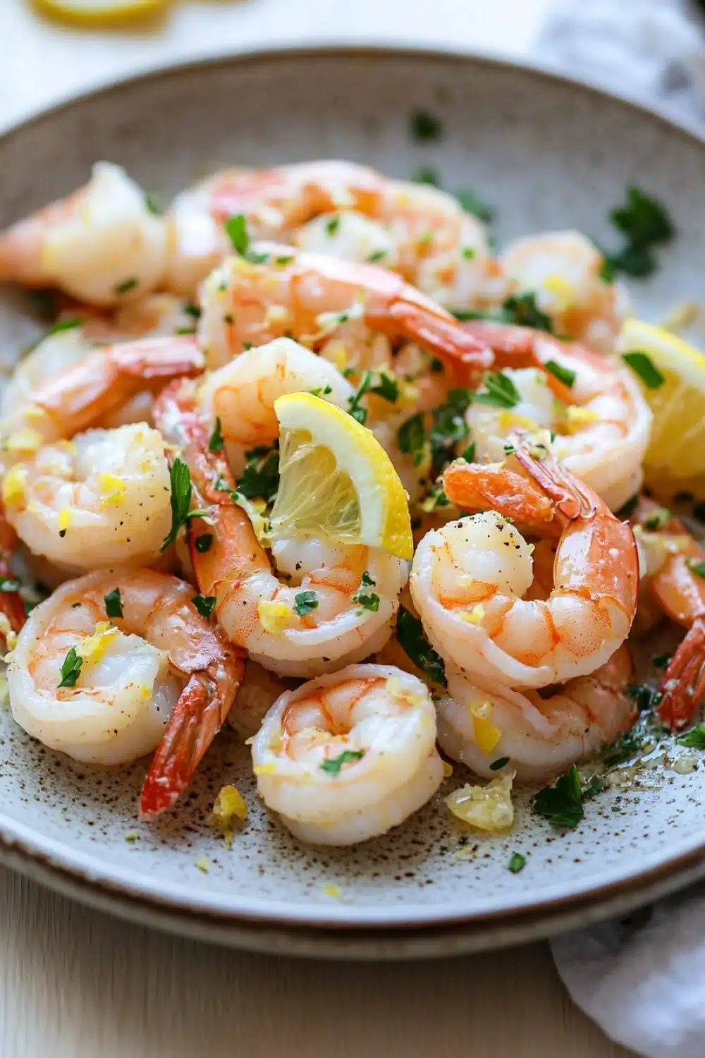 Lemon butter shrimp skillet - the image shows a plate of cooked shrimp on a wooden table. the shrimp are orange in color and appear to be seasoned with herbs and spices. they are arranged in a circular pattern on the plate, with some of them overlapping each other. there are also slices of lemon wedges scattered throughout the dish. the plate is white with a speckled pattern, and there is a white napkin on the side. the background is blurred, but it appears to be a kitchen countertop.