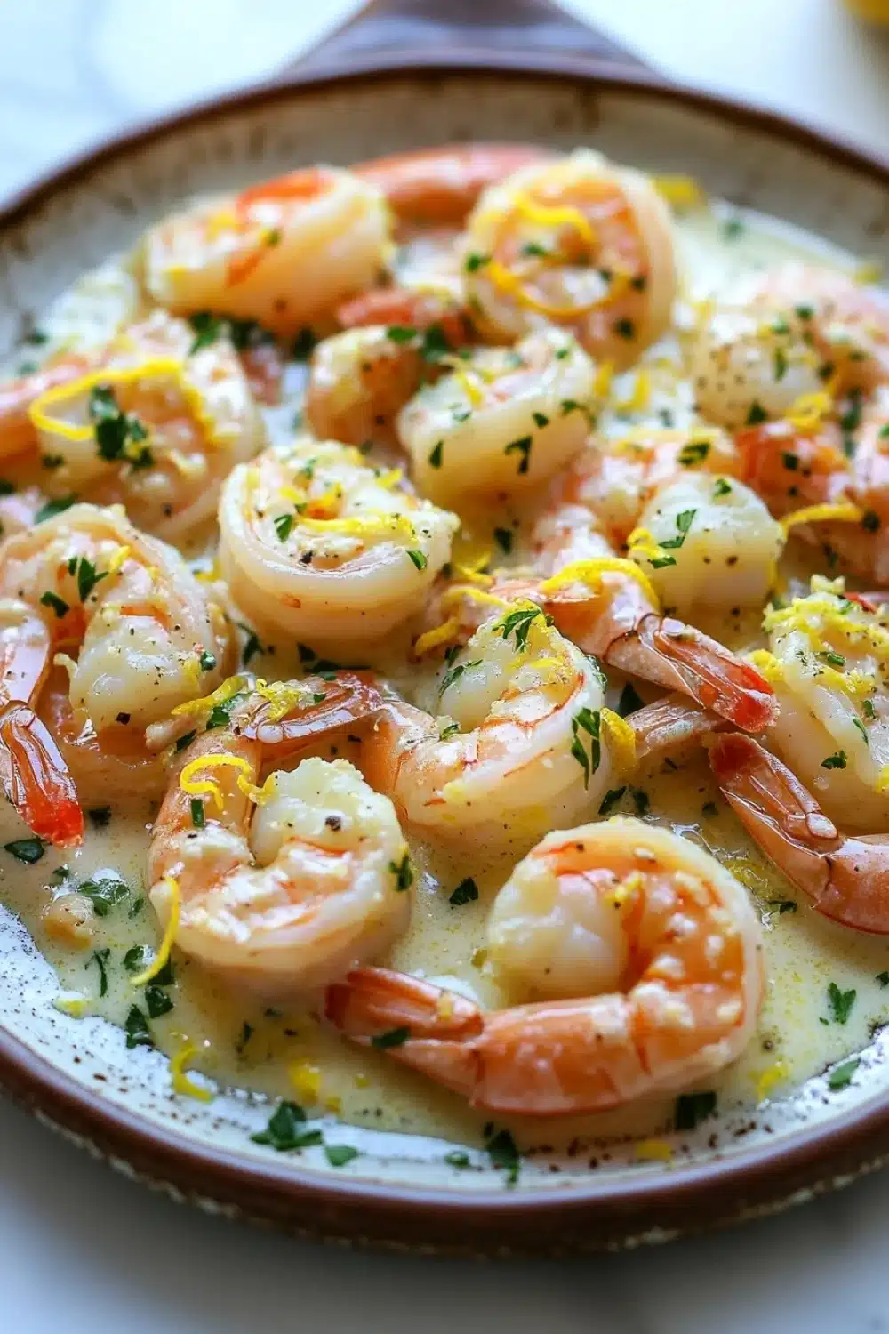 Lemon butter shrimp skillet - the image shows a plate of cooked shrimp in a creamy sauce. the shrimp are pink and appear to be seasoned with herbs and spices. the sauce is a light yellow color and is drizzled over the shrimp. the plate is brown and has a speckled pattern around the edges. the background is blurred, but it appears to be a white tablecloth.