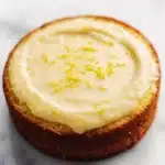 Lemon cake lemon pudding - the image is a close-up of a round cake on a white marble surface. the cake appears to be a lemon cake with a golden brown crust and a creamy white frosting on top. the frosting is spread evenly across the top of the cake and is decorated with small yellow zigzag lines. the background is blurred, making the cake the focal point of the image.