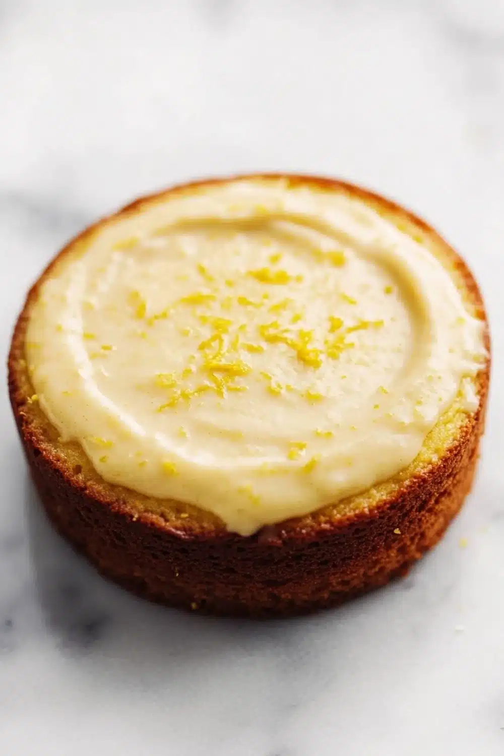 Lemon cake lemon pudding - the image is a close-up of a round cake on a white marble surface. the cake appears to be a lemon cake with a golden brown crust and a creamy white frosting on top. the frosting is spread evenly across the top of the cake and is decorated with small yellow zigzag lines. the background is blurred, making the cake the focal point of the image.