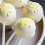 Lemon cake pop design - the image shows a plate of cake pops. the cake pops are round and have a white base with small yellow sprinkles on top. they are arranged in a circular pattern on the plate. the plate is white and has a textured surface. the background is dark, making the cake pops stand out.
