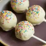 Lemon cake pop design - the image shows four cake pops on a brown plate. the cake pops are round and have a white base with colorful sprinkles on top. the sprinkles are in various colors, including red, blue, green, yellow, and pink. the plate is sitting on a black surface.