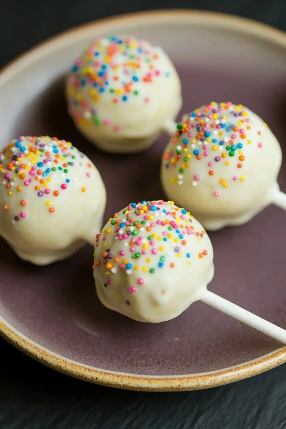 Lemon cake pop design - the image shows four cake pops on a brown plate. the cake pops are round and have a white base with colorful sprinkles on top. the sprinkles are in various colors, including red, blue, green, yellow, and pink. the plate is sitting on a black surface.
