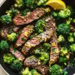 Lemon garlic butter steak and broccoli skillet - the image shows a large black frying pan filled with cooked steak and broccoli. the steak appears to be medium-rare and has a pinkish-brown color. the broccoli is bright green and looks fresh and healthy. there are two lemon wedges on the side of the pan, and a few sprigs of parsley scattered around the pan. the pan is sitting on a wooden table, and there is a gray cloth napkin next to it.
